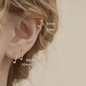 Può includere: Quattro orecchini a cerchio color argento indossati su un orecchio. I due cerchi superiori sono etichettati come 6mm, mentre quelli inferiori sono etichettati come 8mm e 10mm. Gli orecchini presentano un design circolare semplice con una perlina.