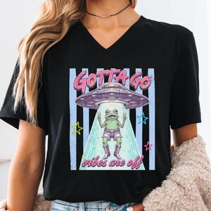 May include: A black V-neck t-shirt featuring a graphic print of a frog wearing shorts and boots, standing under a UFO. The text "GOTTA GO" appears above the UFO, and "vibes are off" is printed below the frog, set against a background of blue stripes.
