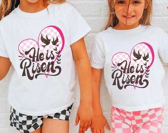 May include: Two white t-shirts with a pink checkered heart design and the text "He is Risen" in brown lettering. The shirts are worn by two children.