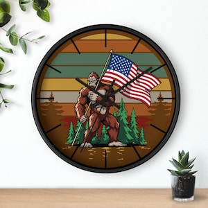 Op de afbeelding: Een ronde wandklok met een zwart frame. De wijzerplaat toont een cartoonillustratie van een Bigfoot met een Amerikaanse vlag. De achtergrond heeft een gestreept ontwerp met bomen en een kleurenschema van zonsondergang.