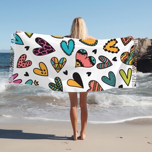 May include: A white beach towel with a colourful heart pattern. The hearts are in various colours, including pink, blue, yellow, green, and black. The towel has a fringed edge.