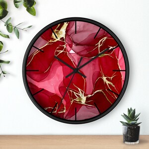 May include: Circular wall clock with a black frame. The clock face displays a striking red and gold abstract pattern. The clock hands are black. The clock is mounted on a white wall.