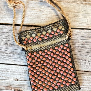May include: A rectangular drawstring pouch featuring a black and gold metallic woven fabric base. The exterior is decorated with rows of small, round, salmon-pink beads arranged in a grid pattern, complemented by a braided tan cord drawstring closure.