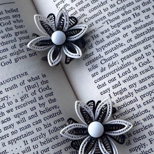 May include: Two black and white flower-shaped brooches with a central white bead are displayed on an open book. The brooches have intricate, layered designs, and the book's pages are filled with text.