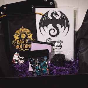 May include: A black box containing purple shredded paper, a black bag with gold text reading "BAG OF HOLDING", a white card with a black dragon illustration, dice, and a small black container. A D&D pin is also visible.