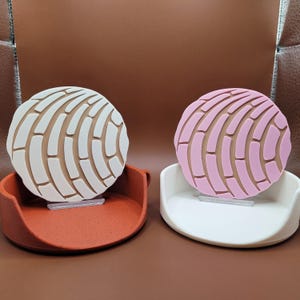 May include: Two phone stands with a concha design. One stand is white with a pink concha and the other stand is brown with a white concha.