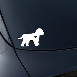 May include: White die-cut vinyl sticker of a dog with a heart detail. The dog is in a standing pose, facing left. The sticker is designed for application on a car or other smooth surface. A simple, clean design.