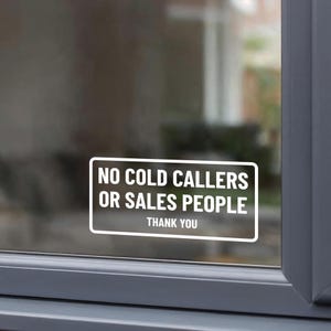 May include: A rectangular white vinyl window decal with rounded corners featuring the bold white text NO COLD CALLERS OR SALES PEOPLE followed by the word THANK YOU in a smaller font size, applied to the surface of a glass window pane.