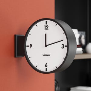 May include: A modern, round wall clock with a black frame and white face. The clock has black hands and numerals, with the brand name "Unikam" printed at the bottom. The clock is mounted on a red wall.