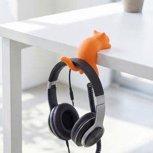 Könnte beinhalten: Orangener Katzen-Kopfhörerhalter an einem weißen Schreibtisch befestigt. Schwarze und silberne Kopfhörer hängen an der Halterung. Im Hintergrund ein kleiner Blumentopf.