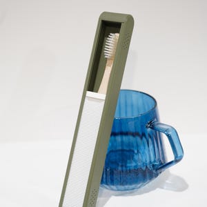 May include: An olive green toothbrush case with a white, ridged front panel, holding a bamboo toothbrush. A blue glass mug with a handle sits behind the case. The items are on a white surface.