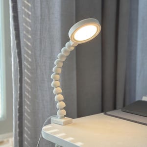 May include: A white desk lamp with a flexible, segmented neck and a round, illuminated head. The lamp is clamped to a white desk, with a gray curtain in the background. The lamp's head is lit, providing bright light.