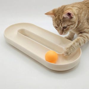 Könnte beinhalten: Ein beiges Katzenspielzeug mit einer ovalen Bahn und einem kleinen orangefarbenen Ball. Eine getigerte Katze interagiert mit dem Spielzeug und greift nach dem Ball. Das Spielzeug ist für die Unterhaltung und Bewegung von Katzen konzipiert.