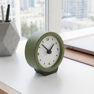 Peut inclure: Une horloge analogique vert olive avec un cadran blanc et des aiguilles noires. L'horloge est posée sur une base noire et affiche l'heure. L'horloge est sur un bureau blanc avec une fenêtre en arrière-plan.