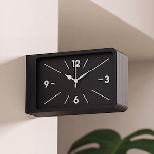 Könnte beinhalten: Eine rechteckige schwarze Wanduhr mit weißen Zeigern und Ziffern. Das Zifferblatt hat ein minimalistisches Design mit Linien, die die Stunden markieren. Die Uhr ist an einer weißen Wand befestigt.
