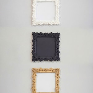 Puede incluir: Tres marcos cuadrados ornamentados en blanco, negro y dorado cuelgan de una pared gris claro. Cada marco tiene un diseño detallado de inspiración floral alrededor de los bordes, con un centro en blanco. Los marcos están espaciados verticalmente.
