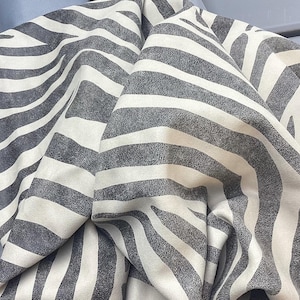May include: A piece of fabric featuring a high-contrast zebra print pattern with dark charcoal gray stripes against an off-white background. The material is draped to create soft folds and shadows, highlighting the textured surface of the textile.