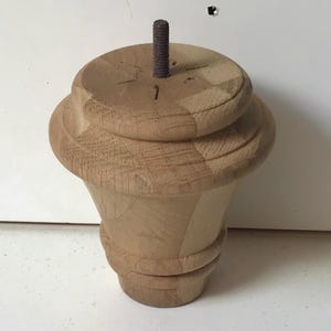 May include: A wooden furniture leg with a turned design. The leg is light brown and has a threaded metal rod extending from the top. The leg has a rounded top and a flared base.