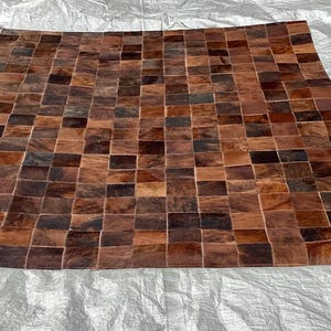 May include: A square rug constructed from a grid of small, rectangular patches of cowhide leather. The patches feature a variety of natural brown, tan, and dark chocolate tones, creating a variegated, multi-colored checkerboard pattern.