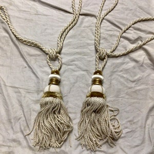 May include: Two white tassel tiebacks with gold accents. The tassels are made of a soft, woven fabric and are attached to a white cord.