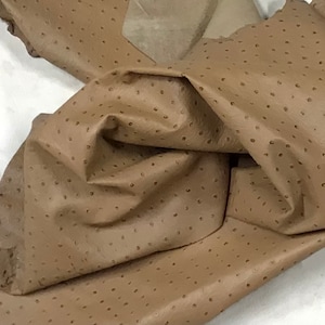 May include: A piece of brown ostrich leather with a textured, dotted pattern. The leather is folded and draped over a white surface.