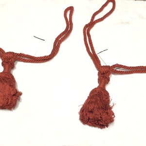 May include: Two rust-colored rope tiebacks with tassels. The tiebacks are made of a thick, braided rope and have a simple knot at the top. The tassels are made of the same rope and are about 3 inches long.