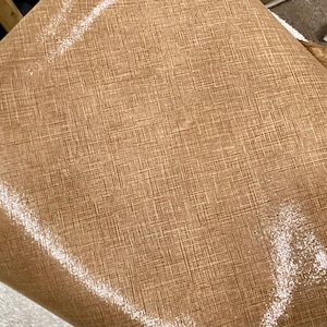 May include: Close-up of a textured, brown fabric with a subtle crosshatch pattern. The material has a slight sheen, reflecting light in a soft, diffused manner. The fabric appears to be folded, revealing its texture and depth.