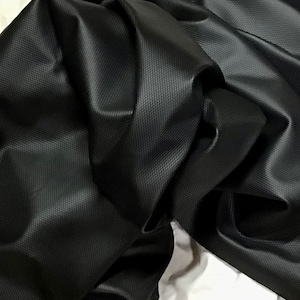 May include: Close-up of a black fabric with a textured, grid-like pattern. The material is folded and draped, creating shadows and highlights that emphasize its texture and sheen. The fabric appears to be a durable material, possibly for upholstery or apparel.