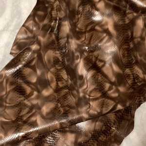 May include: Close-up of a sheet of brown and tan snakeskin leather. The leather has a glossy finish and a detailed pattern of scales. The material is folded, revealing the underside.