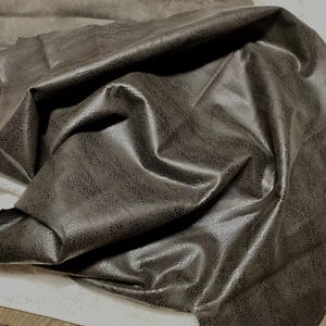 May include: A close-up of a dark gray textured fabric with a subtle snakeskin pattern. The fabric is folded and draped, showcasing its soft and supple texture.