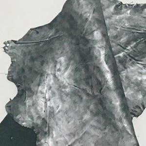 May include: A close-up of a piece of gray leather with a textured surface. The leather is folded and draped, revealing its natural grain and variations in tone. The edges are uneven, suggesting a raw, unfinished state.