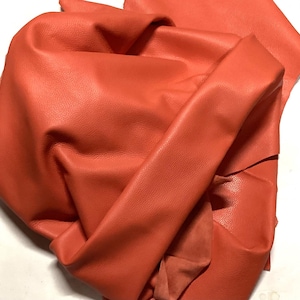 Puede incluir: Primer plano de una vibrante piel de cuero de color coral. El cuero está doblado y texturizado, con grano visible y un acabado mate. El material parece flexible y listo para proyectos de artesanía.