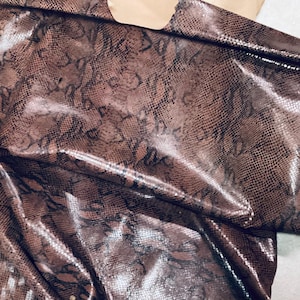 May include: A close-up of a piece of brown and black snakeskin leather. The leather has a glossy finish and a detailed pattern. The material is folded over itself, showing the texture and color variations.