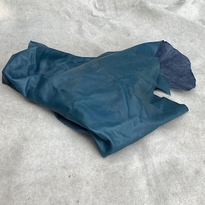 May include: A piece of teal-colored leather with a smooth, matte finish on the top side and a dark, textured, suede-like appearance on the underside where it is folded over. The material is draped in an irregular, soft, and flexible shape.
