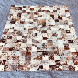 May include: A rectangular area rug constructed from a patchwork of square cowhide tiles. The tiles feature a variety of natural patterns and colors, including shades of brown, white, tan, and dark gray, arranged in a grid-like formation.