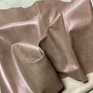 May include: Close-up of a piece of textured, iridescent leather in shades of mauve and tan. The leather has a subtle sheen and a ridged pattern, with folds and creases adding depth. The background is a lighter tan fabric.