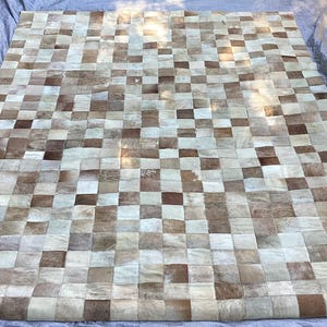 May include: A rectangular area rug constructed from a patchwork of square cowhide tiles in varying shades of cream, beige, tan, and dark brown, arranged in a repeating grid pattern across the entire surface of the floor covering.