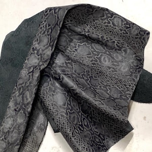 May include: Close-up of a piece of gray snakeskin leather. The leather has a textured pattern and is folded over itself, revealing a dark gray suede backing. The snakeskin pattern is detailed and realistic.