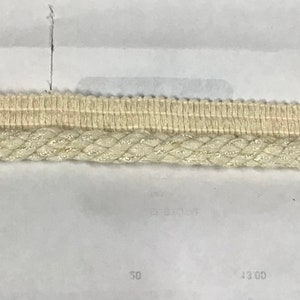 May include: A close-up of two pieces of beige trim. The top piece is a woven fabric with a textured surface. The bottom piece is a braided cord with a subtle shimmer.