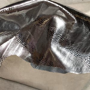 May include: A sheet of silver-colored faux leather with a reptile scale pattern. The material is shiny and reflective, with a textured surface. The underside of the material is a light beige color.