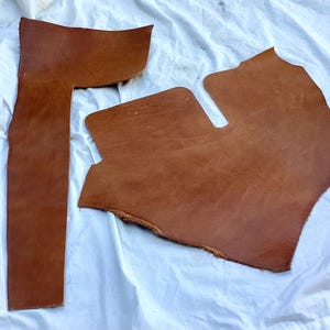 May include: Two irregular pieces of smooth, medium-brown leather with visible natural grain and texture. One piece is long and narrow, while the other is wider with a distinct notch cut out of the top edge, showing raw, unfinished edges.
