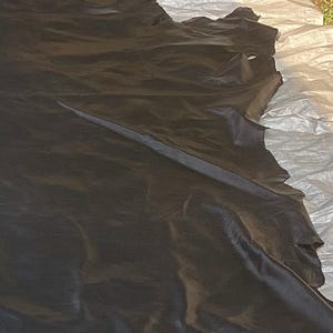 May include: A large, irregular piece of black leather hide with a smooth, matte finish. The material features natural edges and a flexible, draped texture, showing the organic shape of the animal skin with visible folds and creases throughout.
