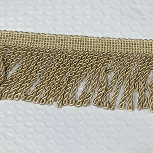 May include: A close-up of a beige fringe trim with a woven texture. The fringe is made of twisted cords and is approximately 1 inch wide.