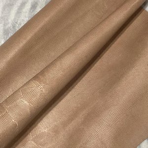 May include: A close-up of a folded piece of tan-colored leather with a textured, scale-like pattern. The leather has a slight sheen and is draped over a soft, white surface. The material appears to be supple and flexible.