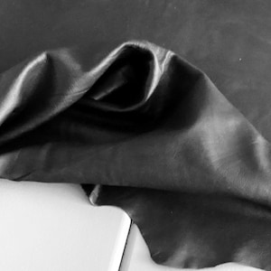 May include: A piece of black leather material with a smooth, matte finish and natural grain texture. The leather is draped to show its flexibility and soft folds, highlighting the deep black color and the subtle variations in the surface texture.