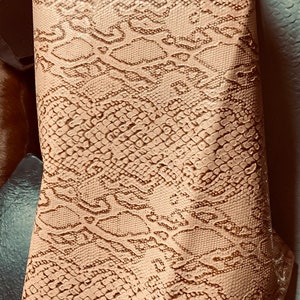 May include: A close-up of a light pink snakeskin-embossed fabric. The fabric has a subtle sheen and a textured surface.