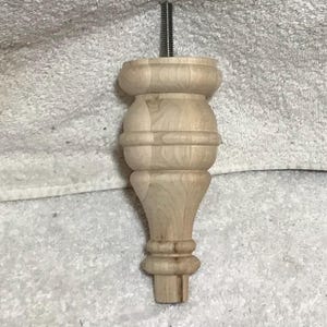 May include: A light-colored, unfinished wooden furniture leg with a threaded metal rod extending from the top. The leg features a turned design with a bulbous upper section, a narrower midsection, and a flared base. The wood grain is visible.