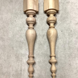 May include: Two unfinished wooden furniture legs. Each leg has a square top, a series of rounded and bulbous sections, and a tapered bottom. The legs are made of light-colored wood and are ready for staining or painting.