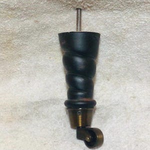 May include: A black, spiral-shaped furniture leg with a brass-colored caster wheel at the bottom. The leg has a threaded bolt at the top for attachment. The caster wheel allows for easy movement.