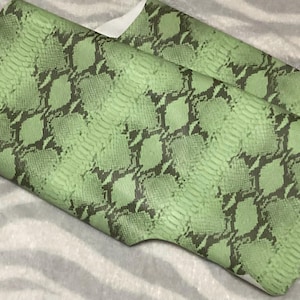 May include: Two pieces of green faux leather with a snakeskin pattern. The pattern features dark grey diamond shapes on a light green background. The material is folded over, revealing a white underside.